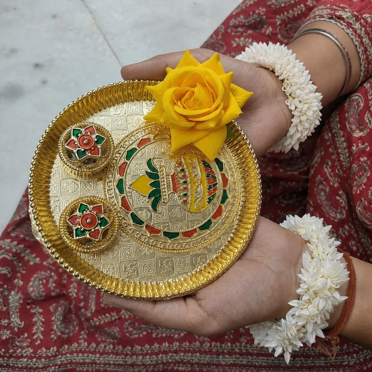 Hands carrying a gold meenakari puja thali with a yellow rose