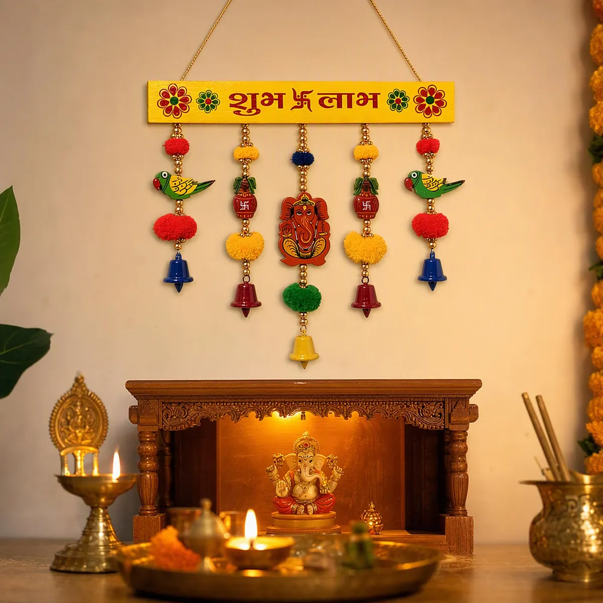 A wooden Shubh Labh toran with parrots and bells above a home mandir lit with diyas
